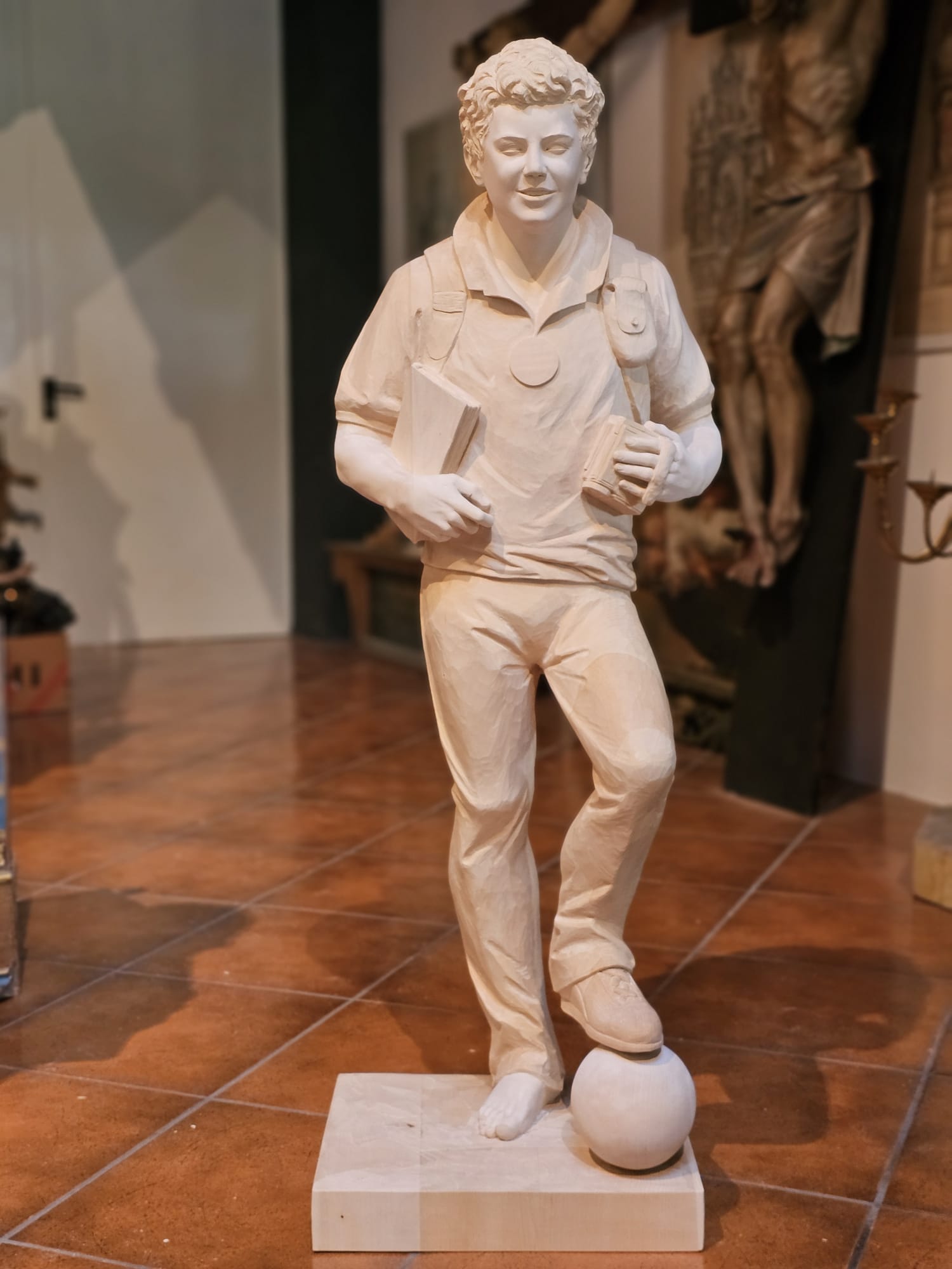 Detailaufnahme der Carlo Acutis Statue im Entstehungsprozess, die ihn mit einem Fuß auf einem Fußball zeigt. In einer Hand hält er ein Buch, in der anderen eine Kamera. Die Statue ist noch unbemalt, wobei die Details der Pose und der Requisiten bereits klar erkennbar sind.